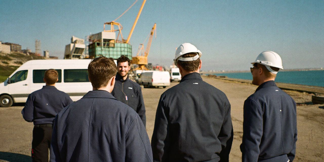 Elektriciens in donkerblauwe werkjassen in gesprek op een bouwplaats in de haven, met kranen en witte bussen