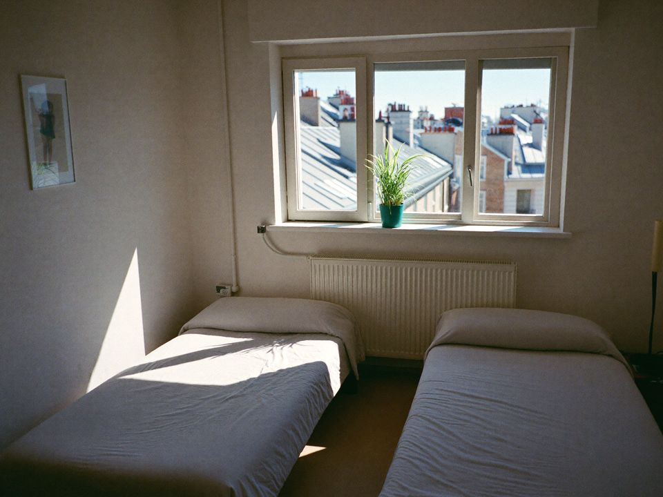 Lichte tweepersoonskamer met twee opgemaakte bedden en een raam met uitzicht op daken